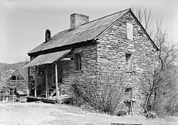 A three story stone building with peaked roof and a front porch to the left built at the crest of a depression to the right