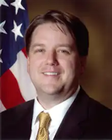 A photographic portrait of a middle-aged man from the shoulders up, wearing a suit, with a necktie, in front of an American flag