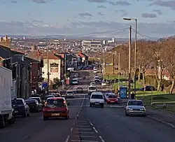 Ina suburban area, a busy main road with vehicles travelling in both directions recedes into the distance. On the left is a row of red-bricked two-storey houses. On the right, an open parkland fringed with mature trees is visible. In the distance is a sunlit landscape of urban and industrial buildings leading to distant, low hills.