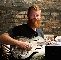 Musician Oliver Anthony, seated on a couch while playing a resonator guitar