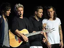 Four young adult males standing next to each other on a stage. Member Niall Horan is holding and playing a guitar. From left to right: Louis Tomlinson, Niall Horan, Liam Payne, and Harry Styles.