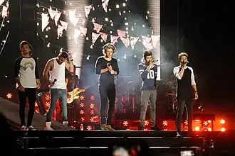 Five young adult males stand next to each other on a stage, all wearing jeans and long-sleeve t-shirts, except for band member Liam Payne, who wears a tank top. From left to right: Louis Tomlinson, Liam Payne, Harry Styles, Zayn Malik, and Niall Horan.