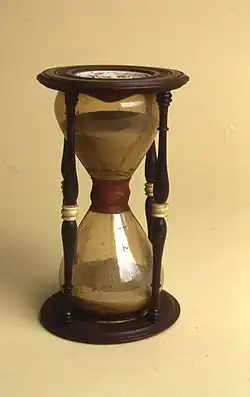 A glass hourglass in a wooden frame, with an ivory joint between the two halves. Sand falls from the top half to the bottom.