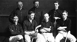 One row of four men seated with three men standing behind. Several have hockey sticks