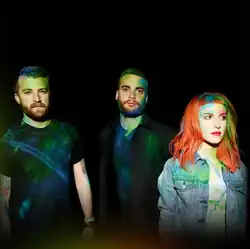 The band members stand on a black background covered in neon paint.