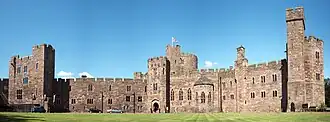 A sandstone castle-like building with a tower slightly to the left of centre. The walls are battlemented and in the foreground is a grassed area