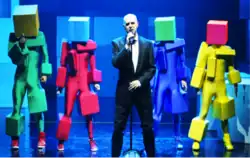 Four dancers wearing colourful cubes on their heads and unitards with cube protrusions stand behind a singer in a black suit