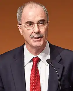 Photograph of Phillip J. Hanlon in a suit and red tie, wearing glasses and speaking at a microphone.