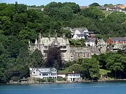 a tree covered landscape with a large mansion and several houses, water in the foreground