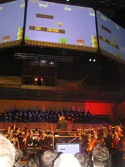 A photograph of a concert of an orchestra performing from the point of view of a member of the audience. Large television screens are featured above the orchestra towards the audience which display gameplay from "The Great Giana Sisters".