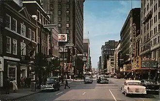 A street in downtown Cleveland in the early 1960s. The building for WJW-TV is to the far left and has a building style that contrasts heavily with the building that surround it, with red brick and white trim, and a cupola which is barely visible at the top of the photo. Among the buildings on the right-hand side are those with more modern exteriors and facades, including marquees for "LOEW'S STATE" and "RKO PALACE".