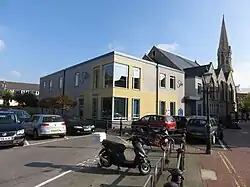 A rectangular grey and pale yellow building, with a church steeple in the background.