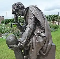 Within a garden sits a bronze statue of Prince Hamlet. He has his right hand on his face as he looks towards a skull that he holds in his left hand. His expression is melancholic.