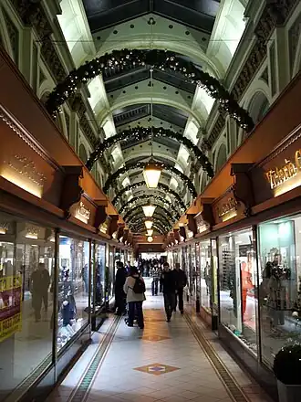 An ornate shopping arcade, lit up by ground floor shop windows, shop signs and fairy lights from above, in the tall ceiling space. The floor is surfaced with white tiles, with linear patterns along the edges of the walkway and diamonds interspersed along the centre.