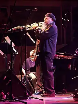 A man on a stage wearing all black and a cap on his head, playing a trumpet into a microphone. Behind him is a man holding a saxophone and another man sitting in a chair. Music stands and additional microphone stands are on the stage in front of them.