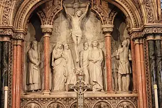 The ornate reredos at the high altar was designed by John Oldrid Scott and sculpted by Mary Grant of Kilgraston in Perthshire - one of the most eminent female sculptors of 19th-century Britain, with numerous commissions from the rich and famous. The reredos is in alabaster with inlaid coloured marbles and depicts the crucifixion scene around the cross at Calvary: to Jesus' left, his mother Mary; and to his right, faithful disciple John. At Jesus' feet is a distressed Mary Magdalene. In addition there are carved wingéd angels atop the structure, and below the crucifixion scene are five marble disks each displaying an angel's face and wings. The figure to the far left of the reredos, in its own outer archway, is St Columba and to the right is St Margaret of Scotland.