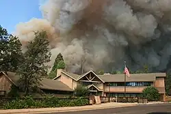 A leaning column of dark brown smoke fills the sky behind a complex of wooden buildings surrounded by trees