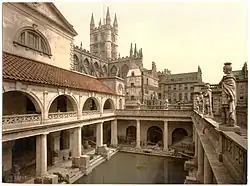 A late-nineteenth-century Photochrom of the Great Bath at the Roman Baths. Pillars tower over the water, and the spires of Bath Abbey – restored in the early sixteenth century – are visible in the background.