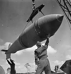 Black and white photo of a large bomb being hoisted. Two men wearing military uniforms are standing below the bomb, and steadying it with their hands.