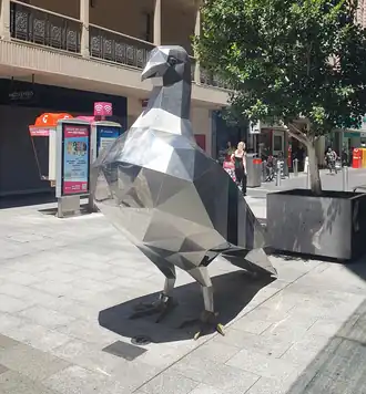 A tall, mirrored stainless steel sculpture of a pigeon in low-poly style shines under the midday sun.