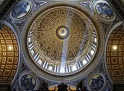 Photo looking up at the dome's interior from below. The dome is decorated at the top with a band of script. Around its base are windows through which the light streams. The decoration is divided by many vertical ribs which are ornamented with golden stars.