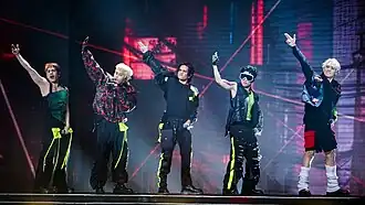 A group photo of SB19 performing onstage, each wearing black clothing with neon highlights; each member has their right arm raised in the air