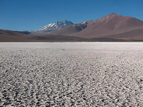 Mountains, including a snow-covered one in the middle, rise above a rough white surface