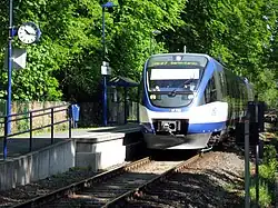 A blue-white three-segment diesel multiple unit on a single track, unelectrified railway line. It is stopping at a small station with one platform featuring a clock, lamps, rubbish bins and shelter, located in a forest.