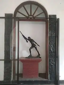 A freedom fighter sculpture with a rifle inside a glass chamber behind a colonial-style door.