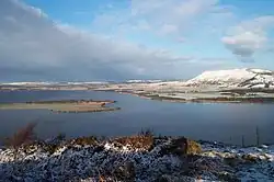 A lake in winter with low snow-covered hills in the background and a brushwood slope in the foreground beneath a mottled blue sky.