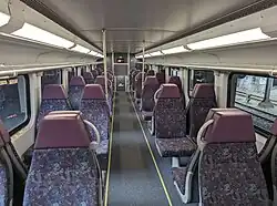 The interior of an empty train car with seats arranged in groups of four—pairs that face inwards—that are separated by an aisle. Large windows and an overhead rack can also be seen.
