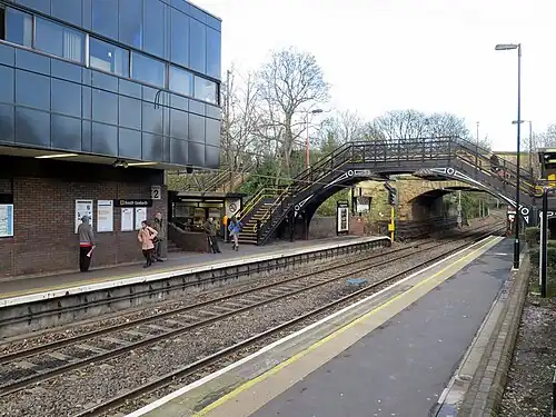 A station with two platforms and tracks. In the foreground is a 19th-century pedestrian bridge.