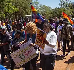 A parade of people walking holding banners and signs. A people car is in the background