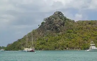 Photo of a body of water with at least four boats anchored in it. Behind the boats is a large rocky hill, covered in vegetation.