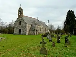 A stone church seen from the southwest, with a slate roof and red tiled ridge. At the west end is a door a window and a double bellcote; protruding from the south side is a porch