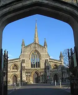 View through an archway of end of a church with a central door flanked by canopied niches containing statues. Arched window above the door and spire behind.