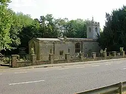 A stone church with a tower on the right. This has a battlemented parapet beyond which is a small pyramidal roof.