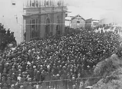 Hundreds of men in black garb and flat-brimmed caps blocking the street, all looking towards a tall building