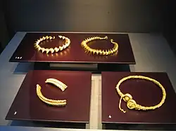 Three elaborate gold coloured torcs lie on dark red surfaces. A fourth torc is in two pieces.
