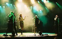 Four singers perform in the spotlights on stage. One is a female in shorts, a gauzy beige skirt and a leather belt, and plays the fiddle, The males are wearing black sleeveless shirts, black pants and have military-looking gear. A rock band is in the background.