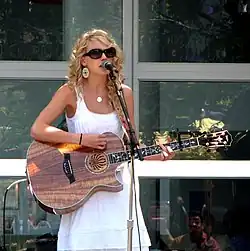 Taylor Swift in sunglasses playing a wooden acoustic guitar and singing onto a mic