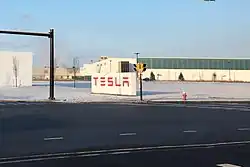 Large white sign, with red letters spelling "Tesla", in the parking lot of a long, low building.