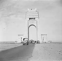 Black and white photograph of the Arch from the front. A paved road can be seen extending from the foreground through the arch, on which several vehicles drive in both directions. A figure can be seen silhouetted at the top of the arch.