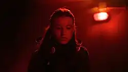 Ellie looks down intensely, surrounded by a red huge from a background light.