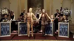 Two female vocalists perform on stage foreground to eight bandmates on the set of Gossip Girl.