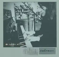 Karen Peris plays piano, with two microphones placed behind her. Behind the microphone is a cabinet full of books, vinyl records, and ornaments. Even further in the distance is a couch and coffee table.