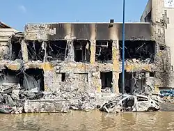 A severely-damaged building, looking as if it were a cross-section. In front is a burned car partially submerged in murky brown water.