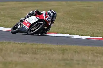 Rider at a left bend on a red and white motorcycle in the middle distance with grass in front and behind track