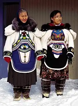 Two Inuit women in clothing covered with detailed beadwork and colorful fabric designs. They have fur ruffs on the hoods of their parkas, cloth skirts beneath their parkas, and fur boots.