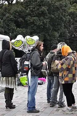 Several people in cold-weather clothing standing before trees visible in the background. The person in the middle of the frame has a few shiny silver heart-shaped balloons attached to their backpack.